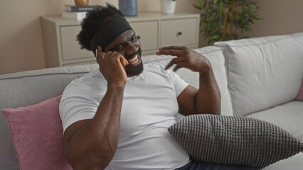 Handsome young african american man with a beard talking on the phone while sitting in a cozy living room of his apartment.