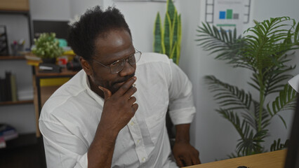 Young african american man with a beard in an office setting, pondering while looking at his computer, with plants and bookshelves in the background.