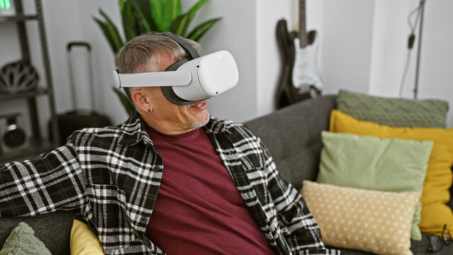 A middle-aged man with gray hair enjoys virtual reality in a cozy living room setting, evoking modern technology and leisure. - Powered by Adobe