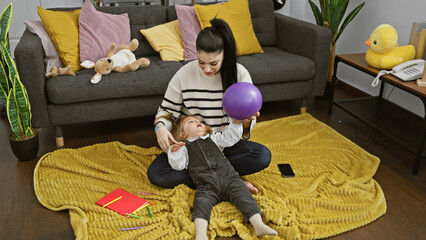 A mother plays with her daughter, holding a purple balloon in a cozy living room with a grey couch and plush toys.