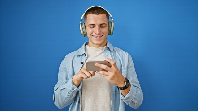 Smiling young hispanic man with headphones using smartphone against a blue wall.