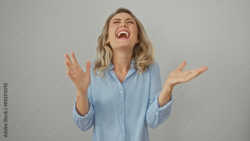 Poster Laughing young blonde woman in a blue shirt with raised hands against a white backdrop.