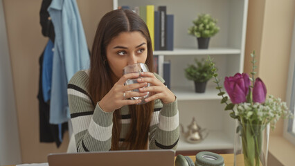 A thoughtful hispanic woman sips water indoors, surrounded by modern home decor and a laptop at her...
