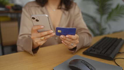 A woman in an office holds a smartphone and credit card while sitting at a desk with a keyboard and mouse.