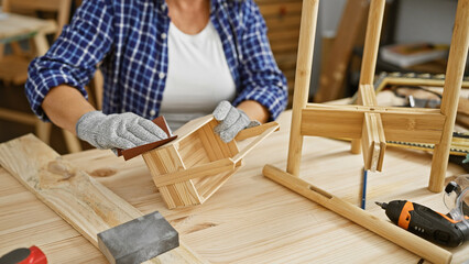 A mature woman sands a wooden chair in her well-equipped carpentry workshop, illustrating craftsmanship and diy.