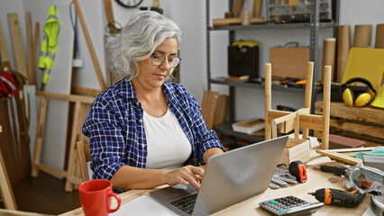 A mature woman in a plaid shirt focused on a laptop in her cluttered woodworking workshop.