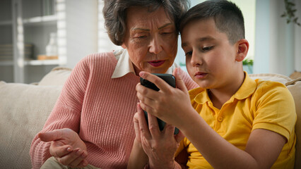 Puzzled elderly woman with phone caring grandson kid boy child teaching grandmother old female use smartphone mobile app shopping in online store at home couch Caucasian family spending time together