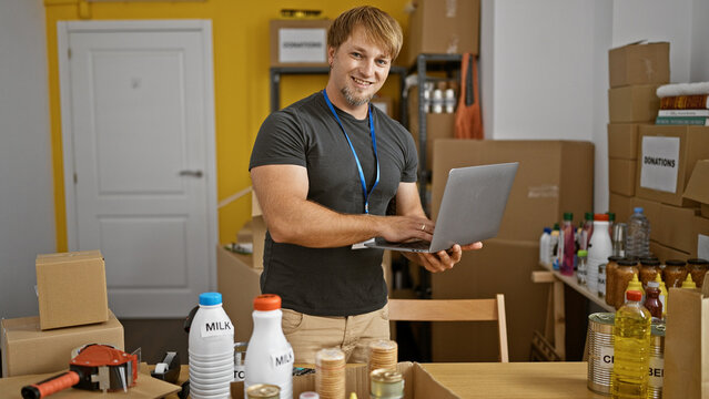 A friendly young man with a beard using a laptop in a storeroom filled with donations and supplies.