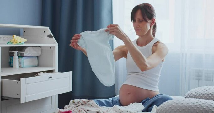 Happy pregnant woman looking at baby clothes with tenderness while sitting on bed in domestic room. Caring future mother making purchases for unborn baby before childbirth