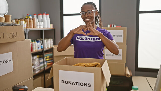 A smiling african american woman makes a heart sign with her hands, wearing a 'volunteer' t-shirt at a donation center.