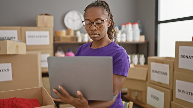 Focused african american woman organizing donations in a warehouse, using a laptop to coordinate the effort.