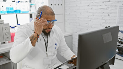 African american man in lab coat working indoors at medical research laboratory with computer and clipboard