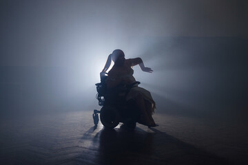 Female silhouette, wheelchair dancer making graceful dance arm movements in a dark space with stage backlit. Inspiration and disability concept.