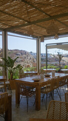 Outdoor restaurant with wicker furniture and scenic mountain view, featuring rustic decor, wooden tables, and ambient hanging lights under a covered patio.