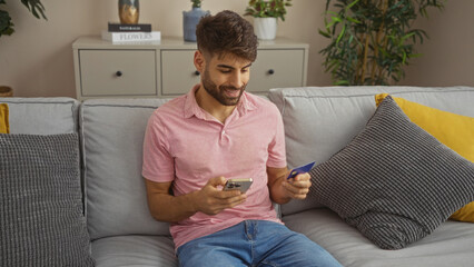 Young man with beard holding phone and credit card while sitting on couch in modern living room indicating online shopping or banking