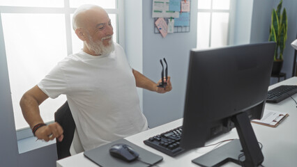 Man with grey hair stretching in an office while holding glasses, mature and senior individual, indoor at a workplace, in front of a computer with a joyful expression