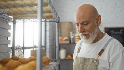 Man with grey hair and beard working in a bakery room, standing near shelves and bread, wearing an apron in an indoor shop interior.