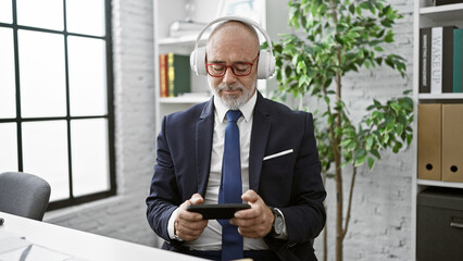 A mature man in a suit enjoys music with headphones in a modern office, holding a tablet.