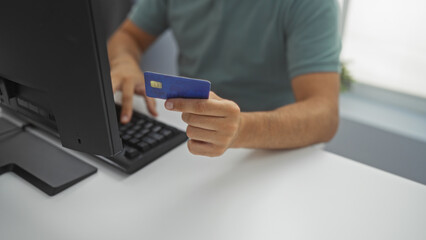 Young man in an office holding a credit card while sitting at a computer, working on an online transaction