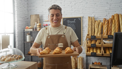 Young man displaying fresh croissants in a cozy bakery interior with shelves of bread and pastries.