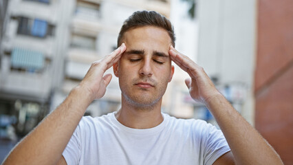 A stressed young hispanic man outdoors in a city, portraying a headache with his fingers on temples.