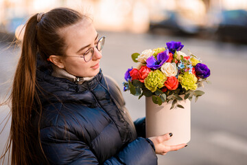 Girl with long hair in a black jacket and glasses stands on a city street with a multi-colored flowers