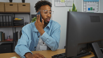 Young african american man working in an office, talking on the phone while looking at a computer screen, with office decor and shelves in the background.