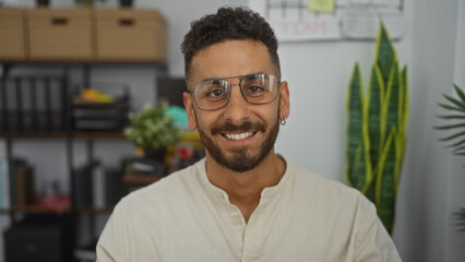 A young, handsome hispanic man with glasses smiles confidently in a modern office, surrounded by plants and shelves.