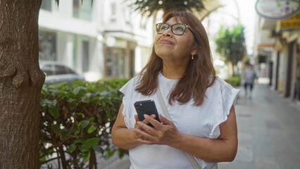 A mature hispanic woman standing on an urban street holding a smartphone, looking up with a thoughtful expression.