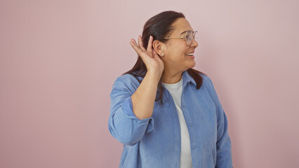 A smiling middle-aged hispanic woman with glasses cupping her ear on a pink isolated background.