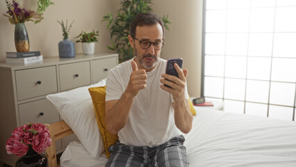 Hispanic middle-aged man conducting a video call on his smartphone while sitting on a bed in a cozy...