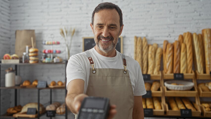 Middle-aged hispanic man working in a bakery shop offering a payment terminal while standing in...