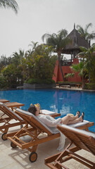 A young woman relaxes on a sun lounger by a resort pool in bali, absorbing the tranquil tropical ambiance.