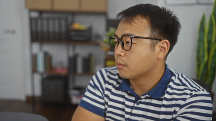 Obraz premium A handsome young chinese man wearing glasses is sitting in an indoor office setting.