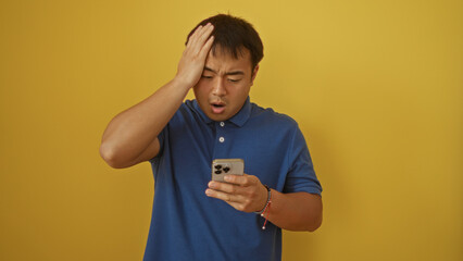 Young chinese man in blue shirt looking at smartphone with shocked expression, isolated against a yellow background.