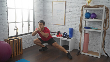 Fototapeta premium A young chinese man performing a squat exercise in an indoor gym with various workout equipment and props in the background.