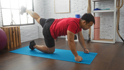 A young chinese man performs a workout on a yoga mat in a gymnasium setting, demonstrating a fitness exercise in an indoor sports center.