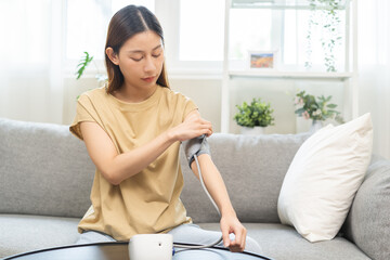 Hypotension problem asian young woman sitting on couch checking high blood pressure and heart rate with digital pressure monitor, making self check up pulse on arm, cuff at home. Healthcare medical.