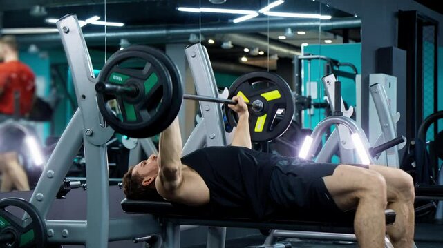 Man in tank top training with barbell in gym. Bench press workout with barbell. Man lifting weights indoor. Chest training. Man is focused and determined. Building muscles indoors. Athlete in tank top