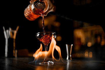 Bartender pours crafted cocktail into a burning glass, close-up spectacle.