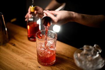 Close-up of female bartender's hands pouring jigger brown liquid into cocktail mixing glass