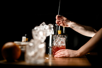Focus on the background where female hands of a bartender stir a brown cocktail with ice