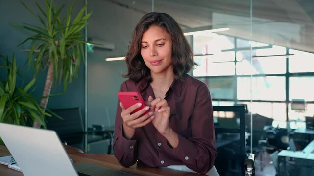 Young Indian business woman manager ceo using cell phone mobile app laptop. Smiling Latin Hispanic mature female businesswoman holding smartphone in office trading, reading, working online. Copy space
