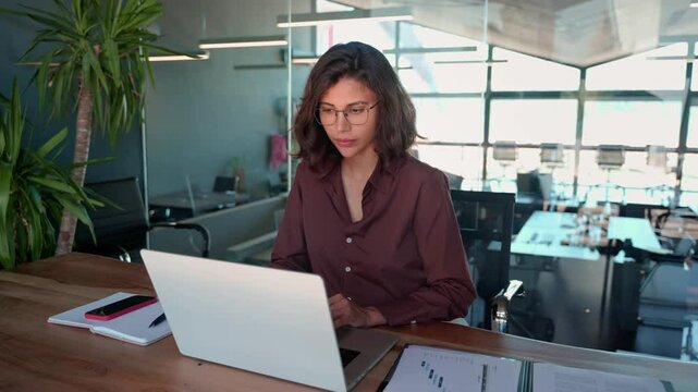 Smiling 30s latin hispanic middle-aged business woman working on laptop computer in modern office. Indian young businesswoman professional employee using pc doing online banking analysing at workplace