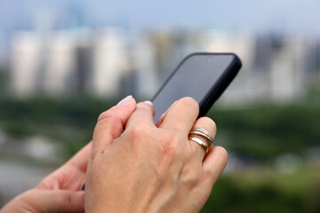 Female hands with smartphone close up on blurred city buildings background. Woman using mobile phone in summer
