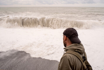 A lonely sad man in a cap and with a backpack looks into the distance at the stormy sea on a cloudy day in autumn