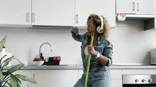 Happy woman with headphones dancing and singing alone in the kitchen using broom as microphone. Craziness and happiness middle age female people having fun cleaning indoor. Leisure activity working