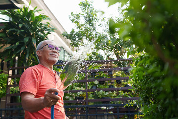 Elderly man in orange shirt watering plants with a hose in his garden, wearing glasses and smiling,...