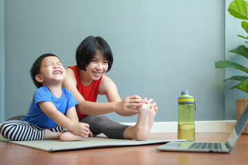 Happy Smiling Asian Mother and little son doing yoga together while watching online class at laptop. Indoor family fitness. Home workout concept