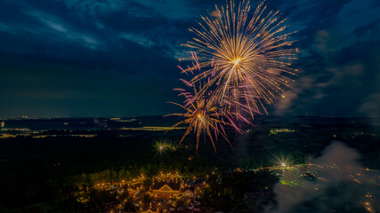 Golden Fireworks Burst Over A Well-Lit Estate, Surrounded By Trees And Fields, With Distant City Lights Adding Depth To The Festive Nighttime Scene.